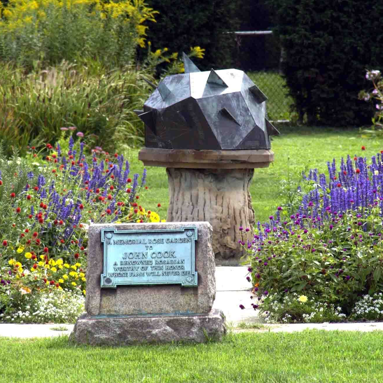 Vibrant flowers surrounding the gnomic block sundial with a dedication to John Cook, renowned rosarian in front.